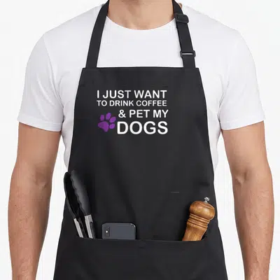 Drink coffee dog apron - White stacked text reading I just want to drink coffee and pet my dogs beside a purple paw — shown on a black cotton canvas apron against a white background