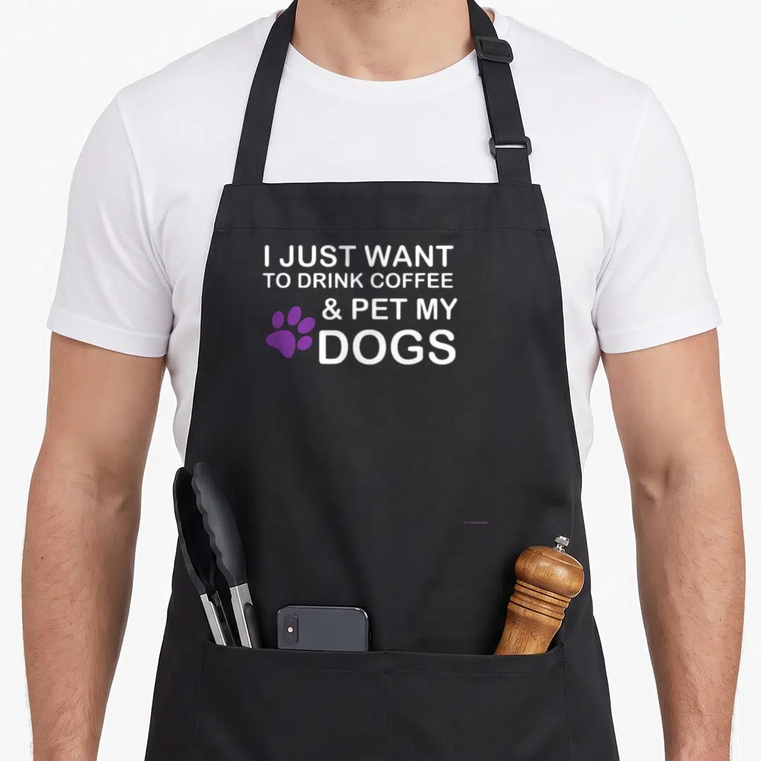 Drink coffee dog apron - White stacked text reading I just want to drink coffee and pet my dogs beside a purple paw — shown on a black cotton canvas apron against a white background