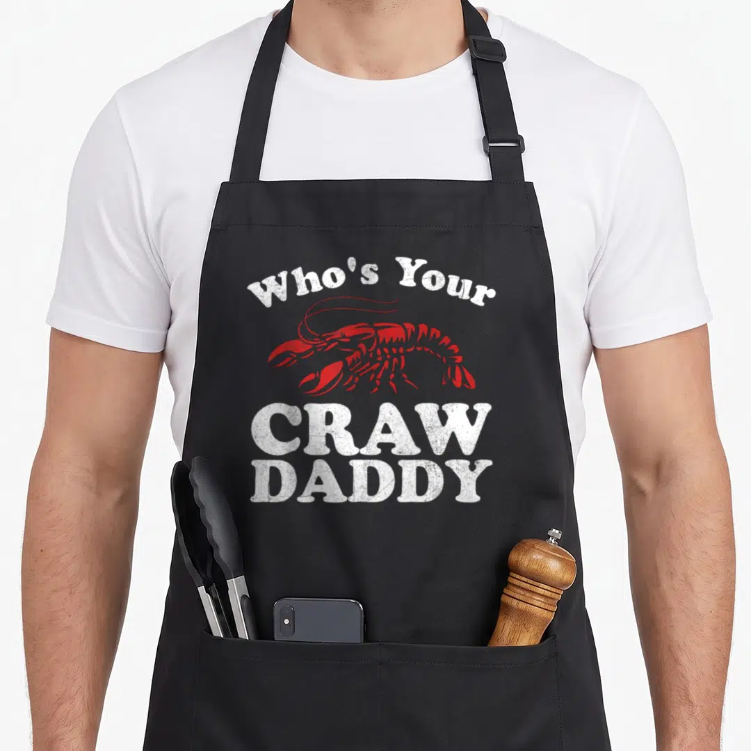 Gras cajun aprons - Red crawfish illustration between arched and stacked white distressed text reading Who's Your CRAW — shown on a black cotton canvas apron against a white background