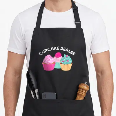 Dealer cupcake apron - Three cupcakes in pink, blue, and yellow wrappers under arched white text reading CUPCAKE DEALER — shown on a black cotton canvas apron against a white background