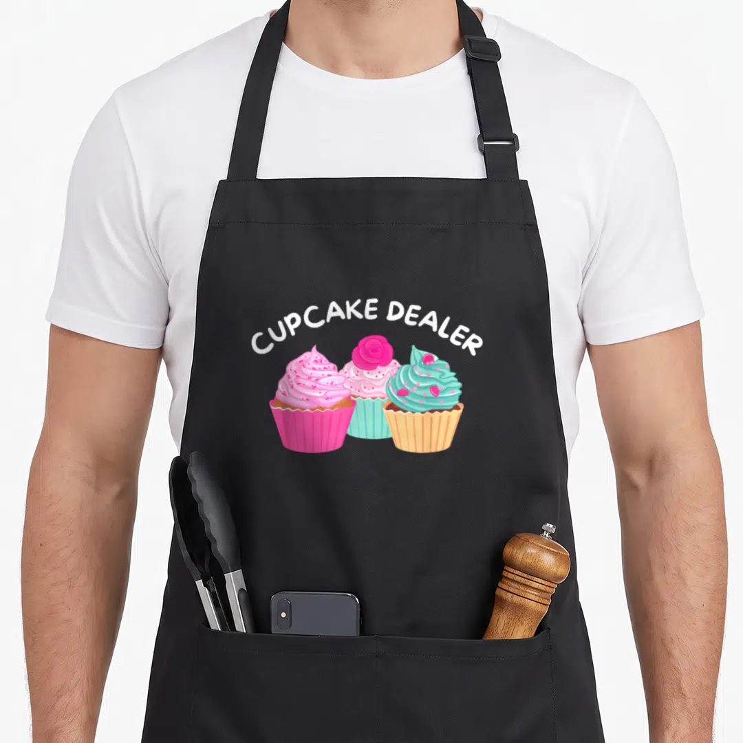 Dealer cupcake apron - Three cupcakes in pink, blue, and yellow wrappers under arched white text reading CUPCAKE DEALER — shown on a black cotton canvas apron against a white background