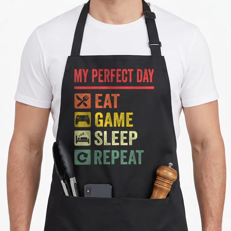 Funny perfect video game apron - Red, yellow, beige, green squares with Eat Game Sleep Repeat text and utensil — shown on a black cotton canvas apron against a white background