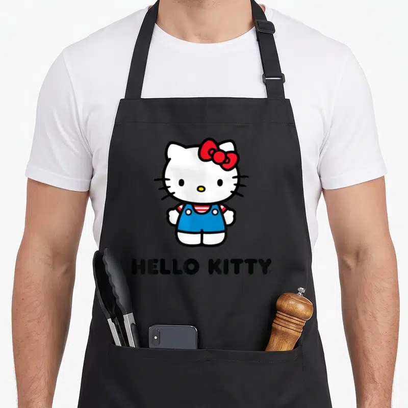 General character aprons - Cartoon Hello Kitty in blue overalls with red bow and black text below — shown on a black cotton canvas apron against a white background