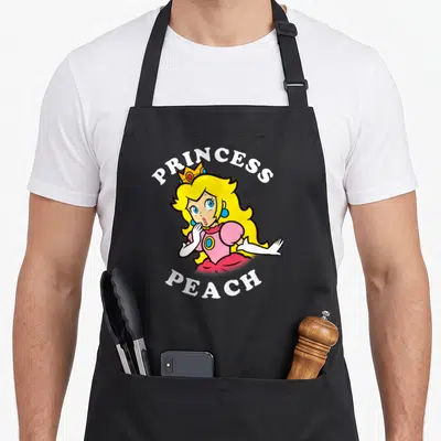 Character princess apron - Princess Peach cartoon portrait with arched PRINCESS PEACH text — shown on a black cotton canvas apron against a white background