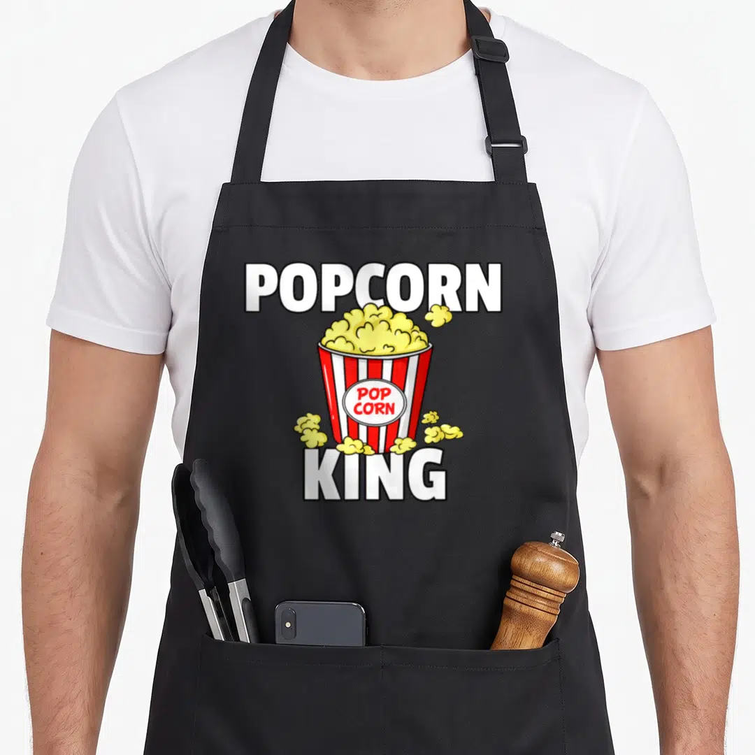Popcorn king apron - Cartoon red white striped bucket with yellow popcorn and POPCORN KING text — shown on a black cotton canvas apron against a white background