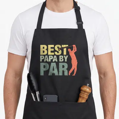 Par papa apron - Best Papa By Par text with orange-red golfer silhouette and distressed yellow green lettering — shown on a black cotton canvas apron against a white background