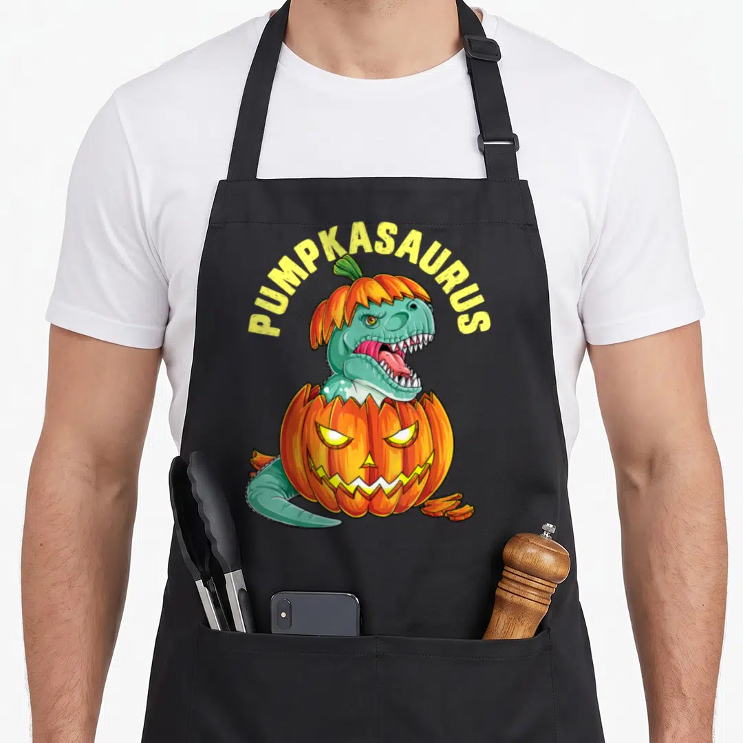 Pumpkasaurus pumpkin apron - Teal dinosaur wearing pumpkin hat emerging from carved jack-o-lantern with arched yellow — shown on a black cotton canvas apron against a white background