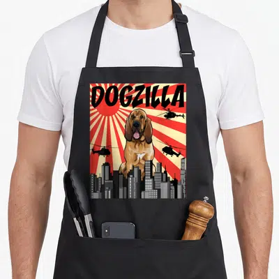 Japanese bloodhound apron - DOGZILLA text over giant brown dog, helicopters, city skyline, red beige sunburst — shown on a black cotton canvas apron against a white background