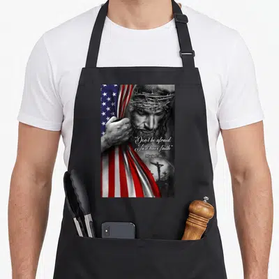Afraid funny christian aprons - Jesus with crown of thorns holding red white and blue flag with scripture text — shown on a black cotton canvas apron against a white background