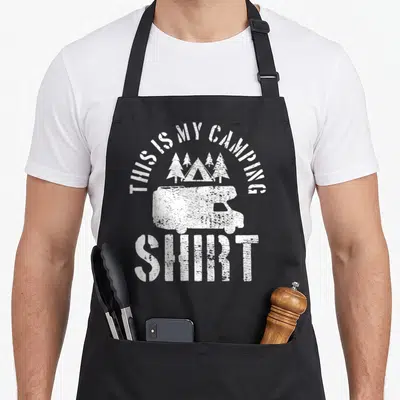 Camping trailer apron - White distressed camper van with tent and four pine trees under arched text — shown on a black cotton canvas apron against a white background