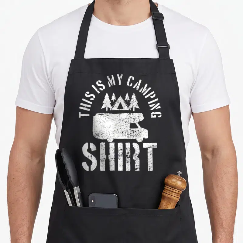 Camping trailer apron - White distressed camper van with tent and four pine trees under arched text — shown on a black cotton canvas apron against a white background