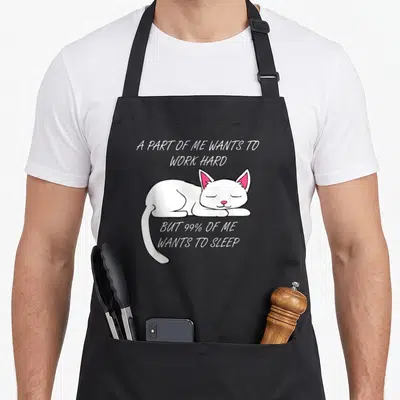 Office kitty cat apron - Sleeping white cartoon cat with pink ears and text about wanting to sleep instead of working — shown on a black cotton canvas apron against a white background