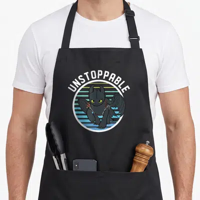 Unstoppable dragon apron - Cartoon black dragon in striped circle with arched UNSTOPPABLE text — shown on a black cotton canvas apron against a white background