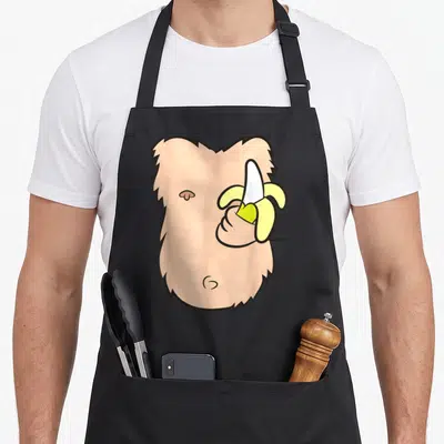 Costume monkey mom apron - Cartoon peach monkey face with hand holding peeled yellow banana — shown on a black cotton canvas apron against a white background