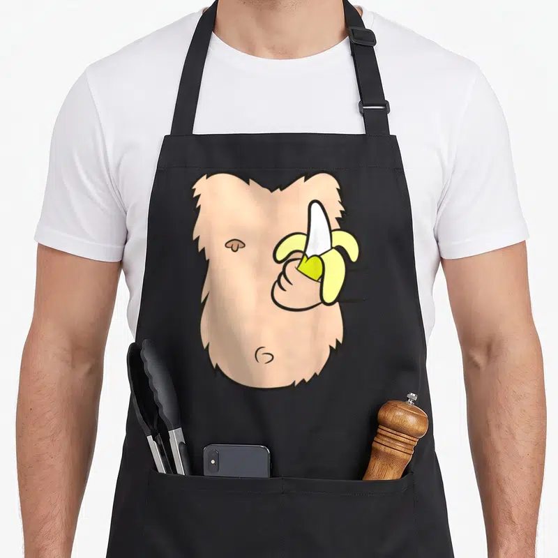 Costume monkey mom apron - Cartoon peach monkey face with hand holding peeled yellow banana — shown on a black cotton canvas apron against a white background