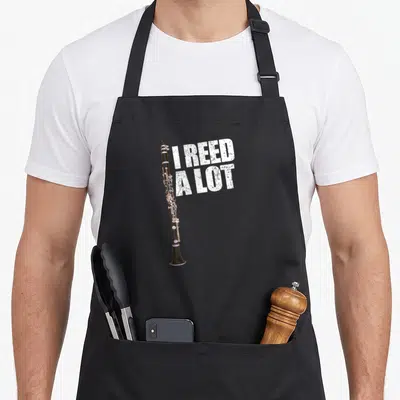Pun lot of aprons - Vertical black clarinet with white distressed text I REED A LOT stacked to the right — shown on a black cotton canvas apron against a white background