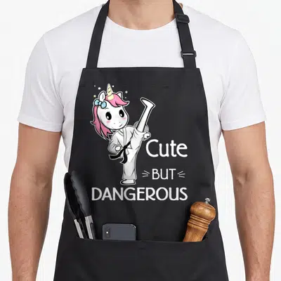 Dangerous unicorn apron - Cartoon unicorn in karate gi kicking next to stacked cute but dangerous text — shown on a black cotton canvas apron against a white background