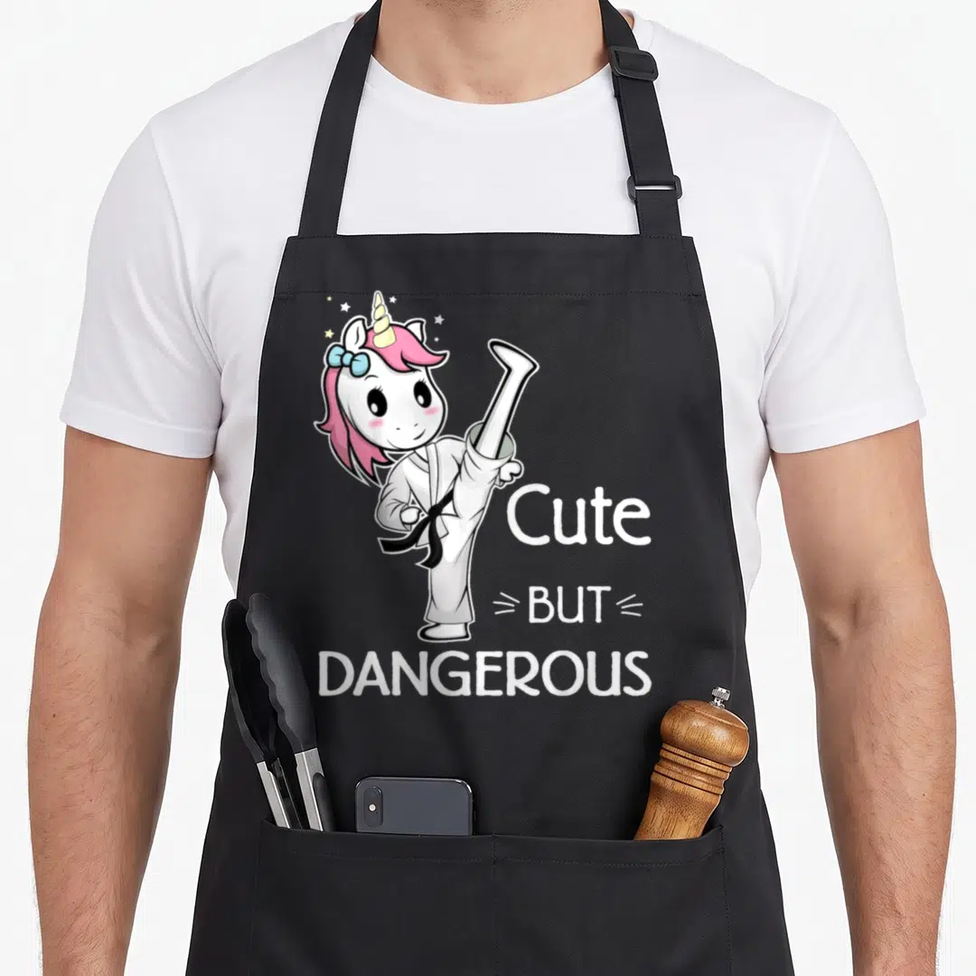 Dangerous unicorn apron - Cartoon unicorn in karate gi kicking next to stacked cute but dangerous text — shown on a black cotton canvas apron against a white background