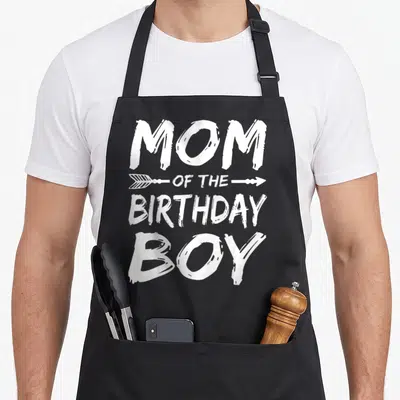 Cute birthday family cooking aprons - White MOM OF THE BIRTHDAY BOY text with arrows on black apron — shown on a black cotton canvas apron against a white background