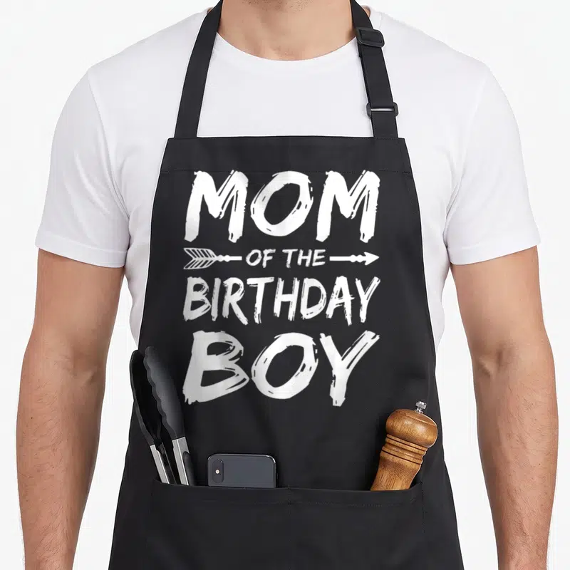 Cute birthday family cooking aprons - White MOM OF THE BIRTHDAY BOY text with arrows on black apron — shown on a black cotton canvas apron against a white background