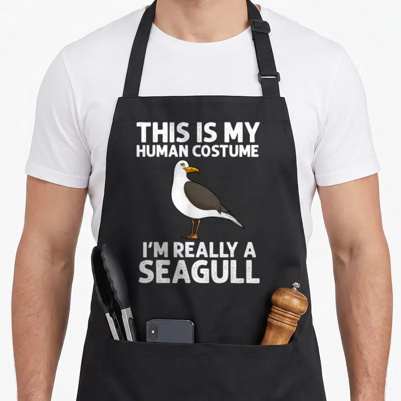 Seabird funny seagull apron - Cartoon seagull between text THIS IS MY HUMAN COSTUME I'M REALLY A SEAGULL — shown on a black cotton canvas apron against a white background