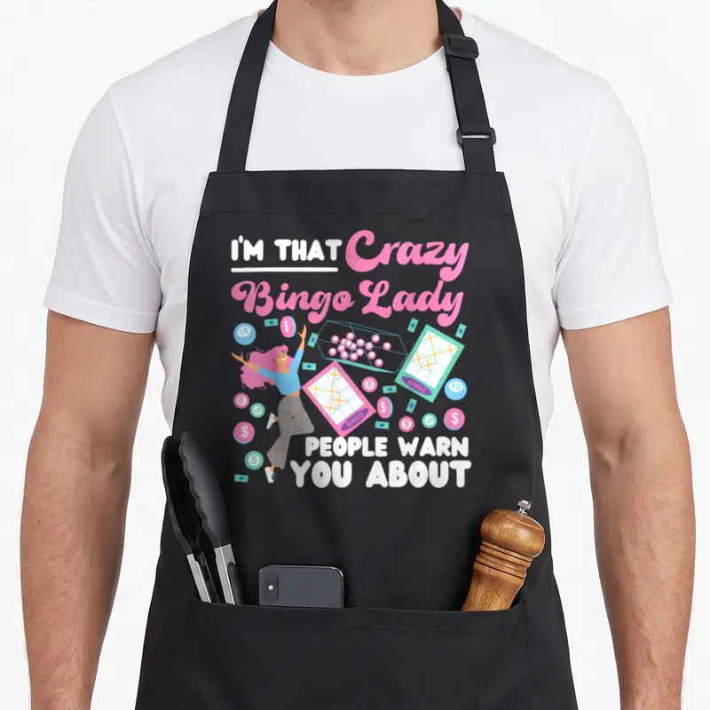 Player warn bingo apron - Cartoon woman surrounded by bingo cards coins and money with pink and white text — shown on a black cotton canvas apron against a white background