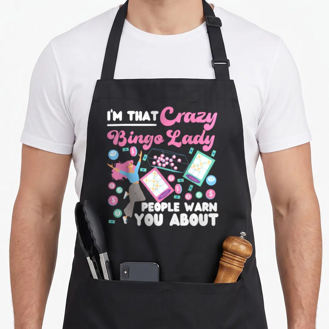 Player warn bingo apron - Cartoon woman surrounded by bingo cards coins and money with pink and white text — shown on a black cotton canvas apron against a white background