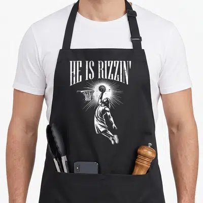 Rizzin easter apron - White distressed HE IS RIZZIN text above jumping basketball player with sunburst — shown on a black cotton canvas apron against a white background