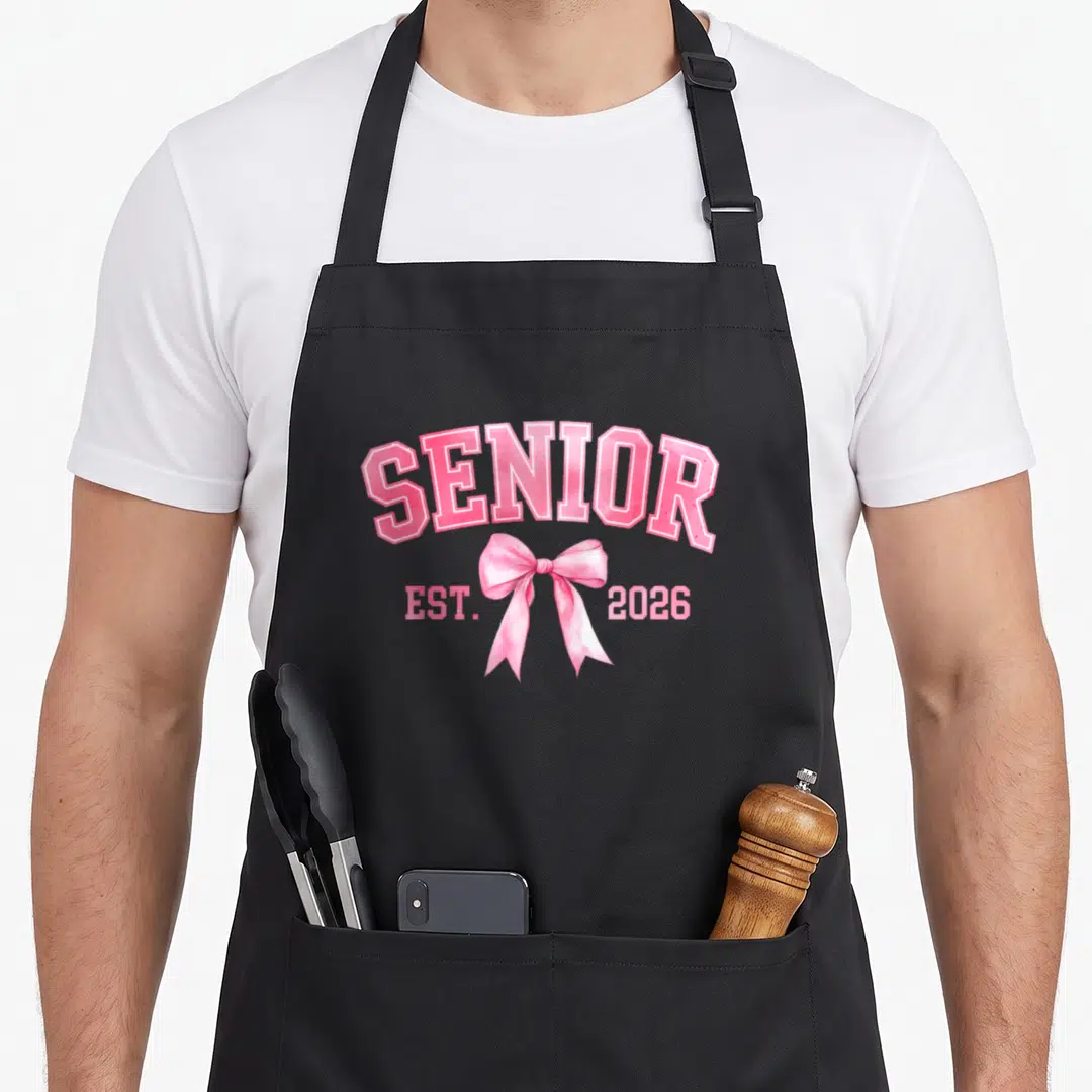Graduation bow apron - Arched SENIOR text over a pink bow with EST. 2026 flanking the ribbon tails — shown on a black cotton canvas apron against a white background