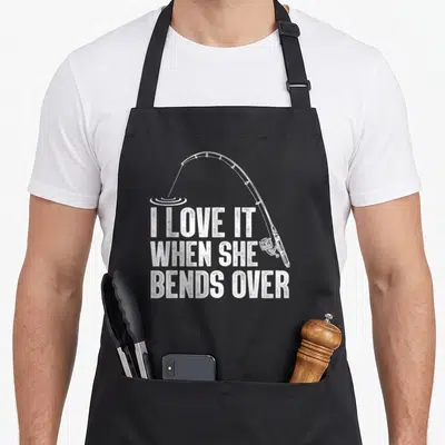 Fisherman rod fishing aprons - White distressed text I LOVE IT WHEN SHE BENDS OVER with bent fishing rod and water — shown on a black cotton canvas apron against a white background