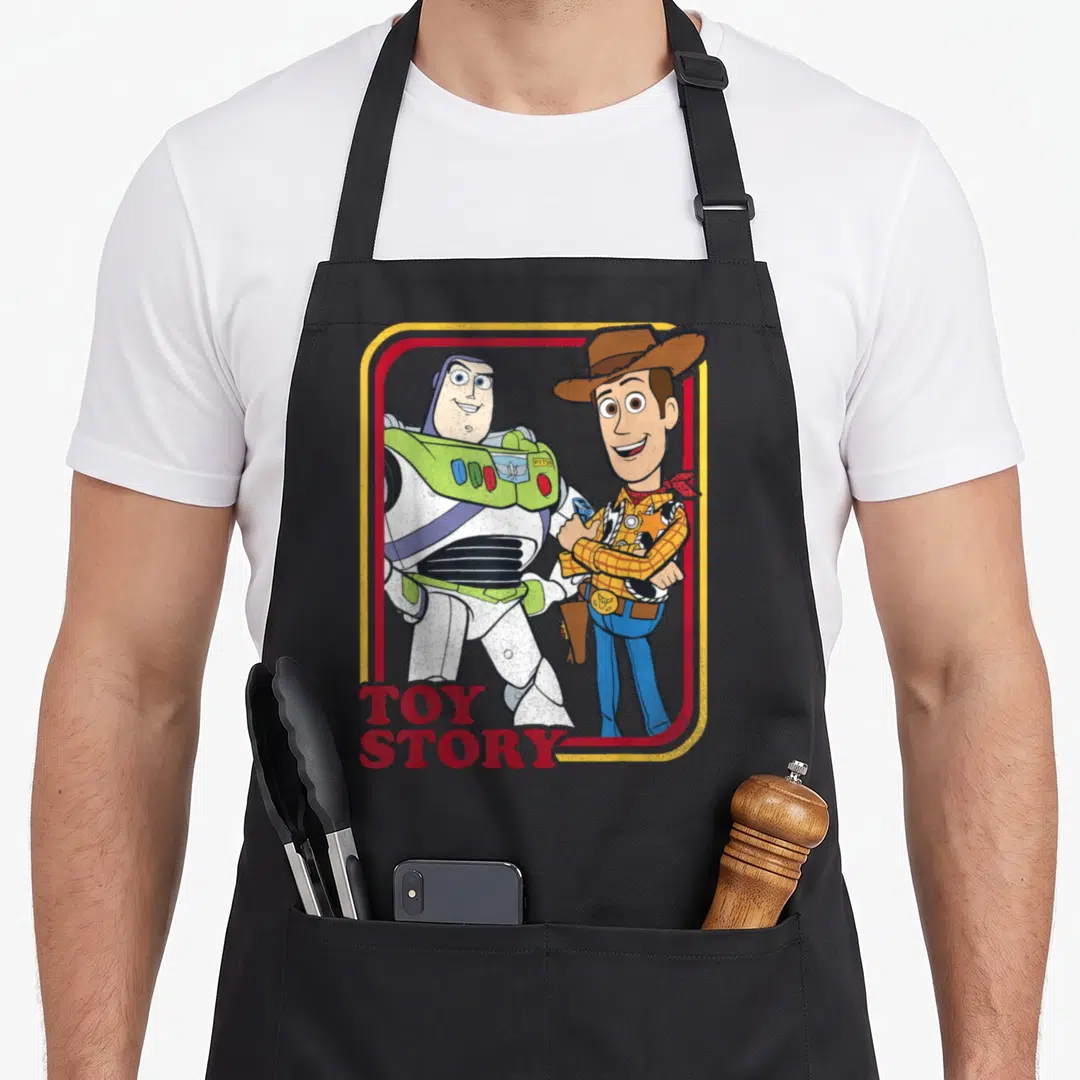 Woody camping apron - Cartoon Buzz Lightyear and Woody standing in red yellow frame with TOY STORY text — shown on a black cotton canvas apron against a white background