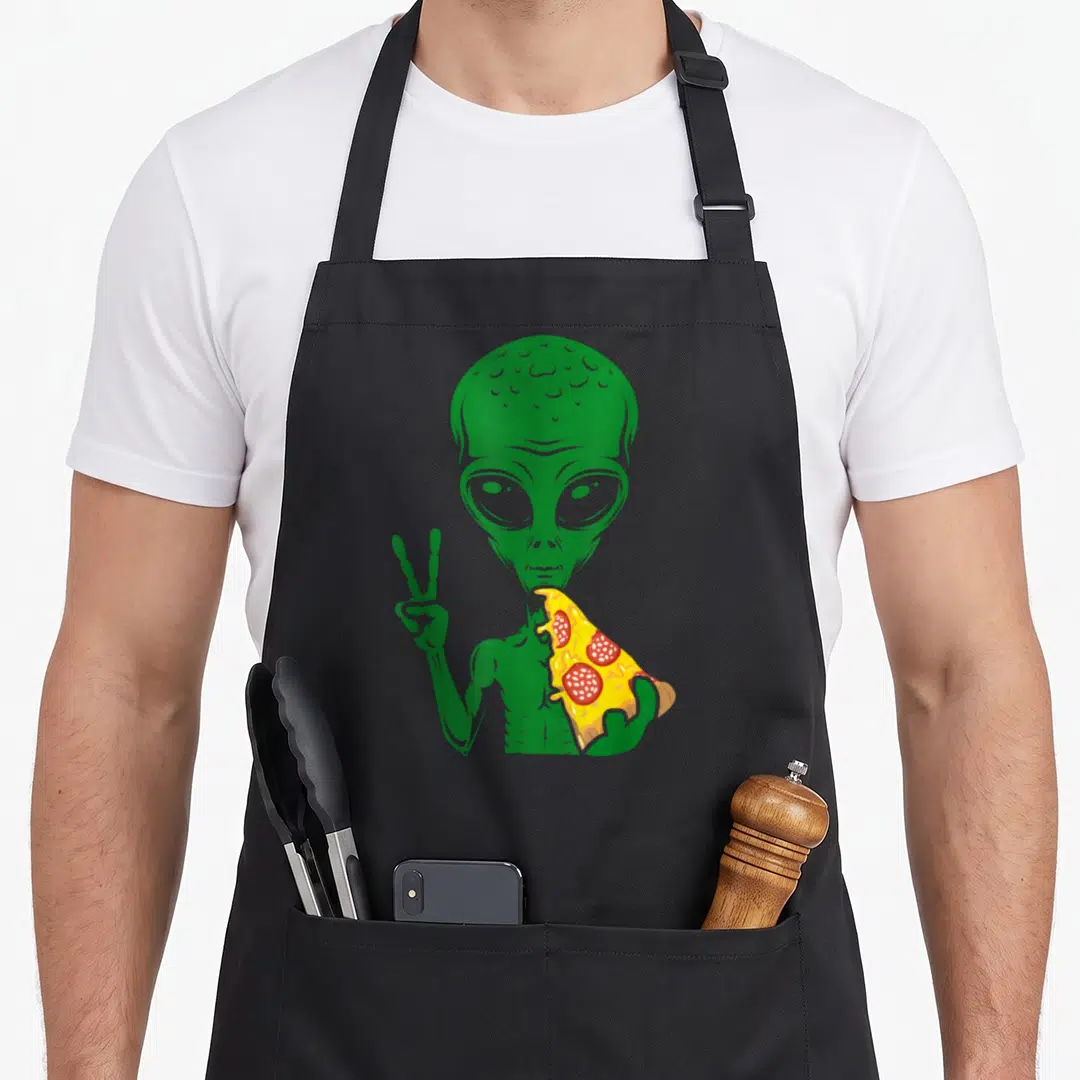 Alien pizza apron - Green cartoon alien holding a yellow pizza slice with red toppings and making a peace sign — shown on a black cotton canvas apron against a white background