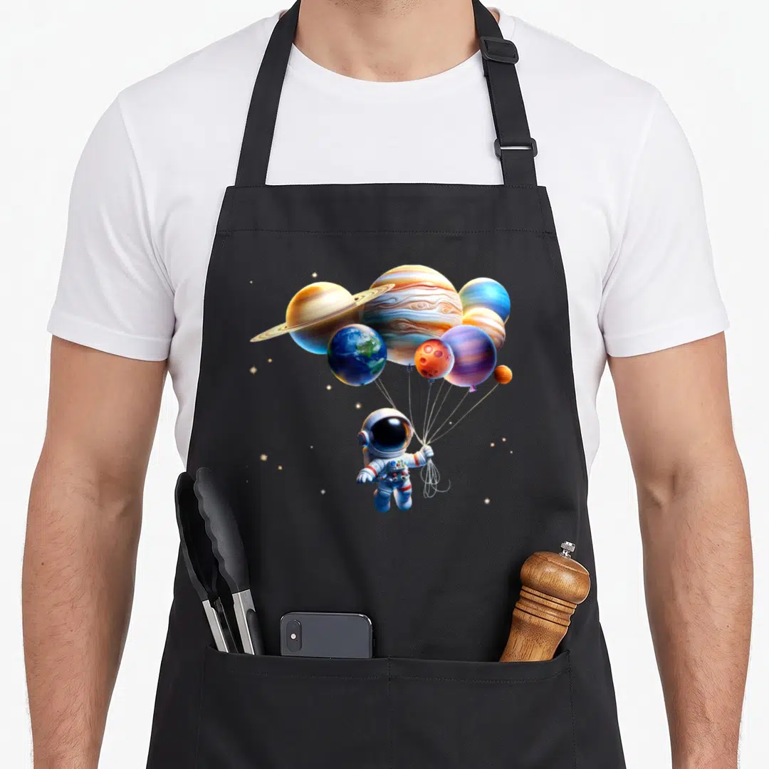 Planet astronaut space apron - Cartoon astronaut holding planet balloons in white blue orange and yellow — shown on a black cotton canvas apron against a white background