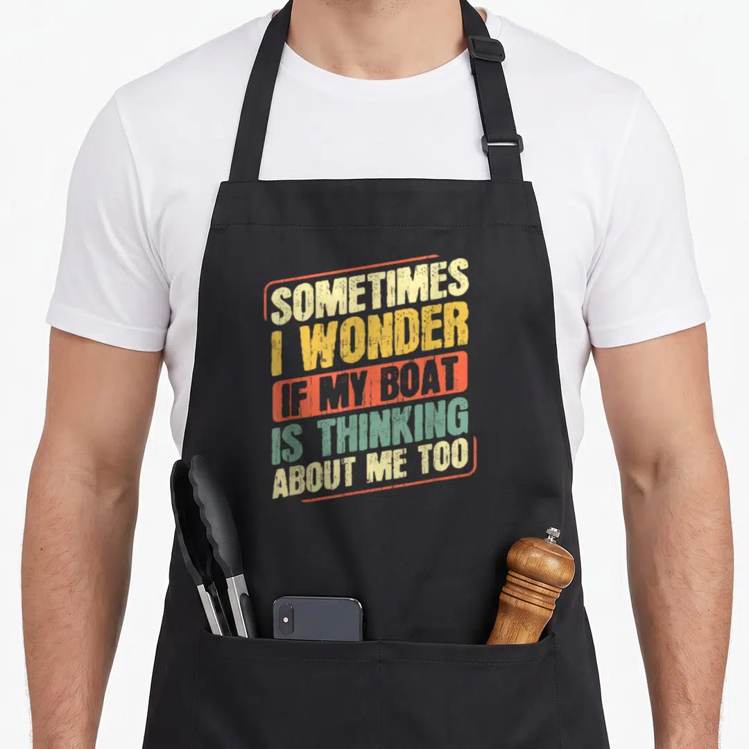 Sometime fishing aprons - Stacked text SOMETIMES I WONDER IF MY BOAT IS THINKING ABOUT ME TOO in beige yellow orange — shown on a black cotton canvas apron against a white background