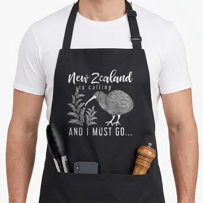 Must new zealand apron - White distressed text New Zealand is calling AND I MUST GO around a standing kiwi bird — shown on a black cotton canvas apron against a white background