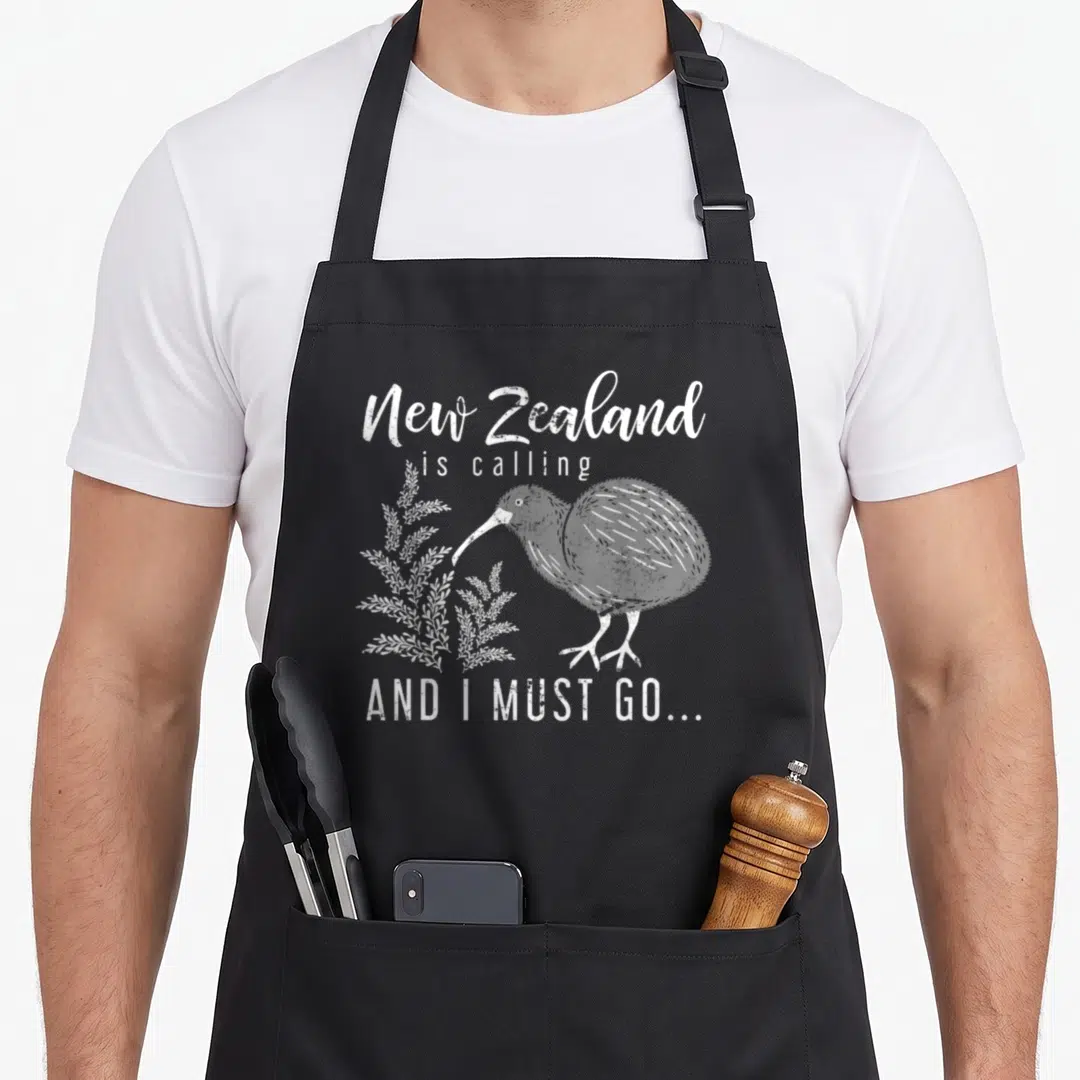 Must new zealand apron - White distressed text New Zealand is calling AND I MUST GO around a standing kiwi bird — shown on a black cotton canvas apron against a white background