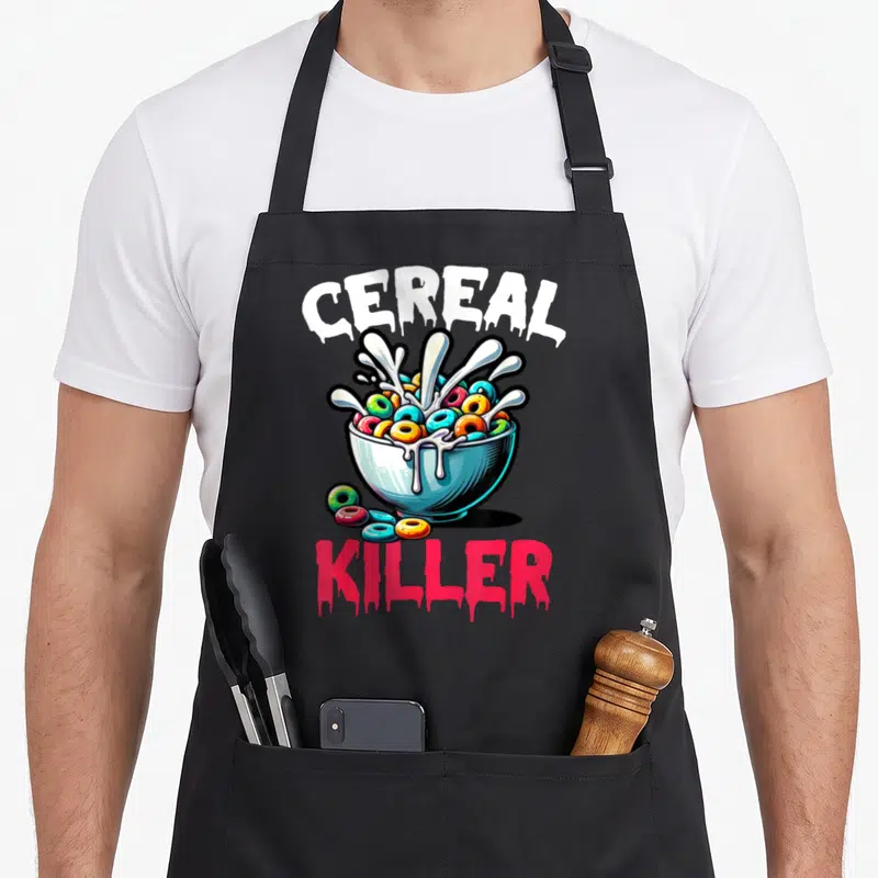 Funny killer cereal apron - Dripping CEREAL KILLER text above and below a bowl of colorful ring cereal with milk — shown on a black cotton canvas apron against a white background