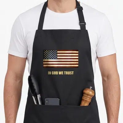 Trust 4th of july apron - Realistic distressed American flag above gold IN GOD WE TRUST text — shown on a black cotton canvas apron against a white background