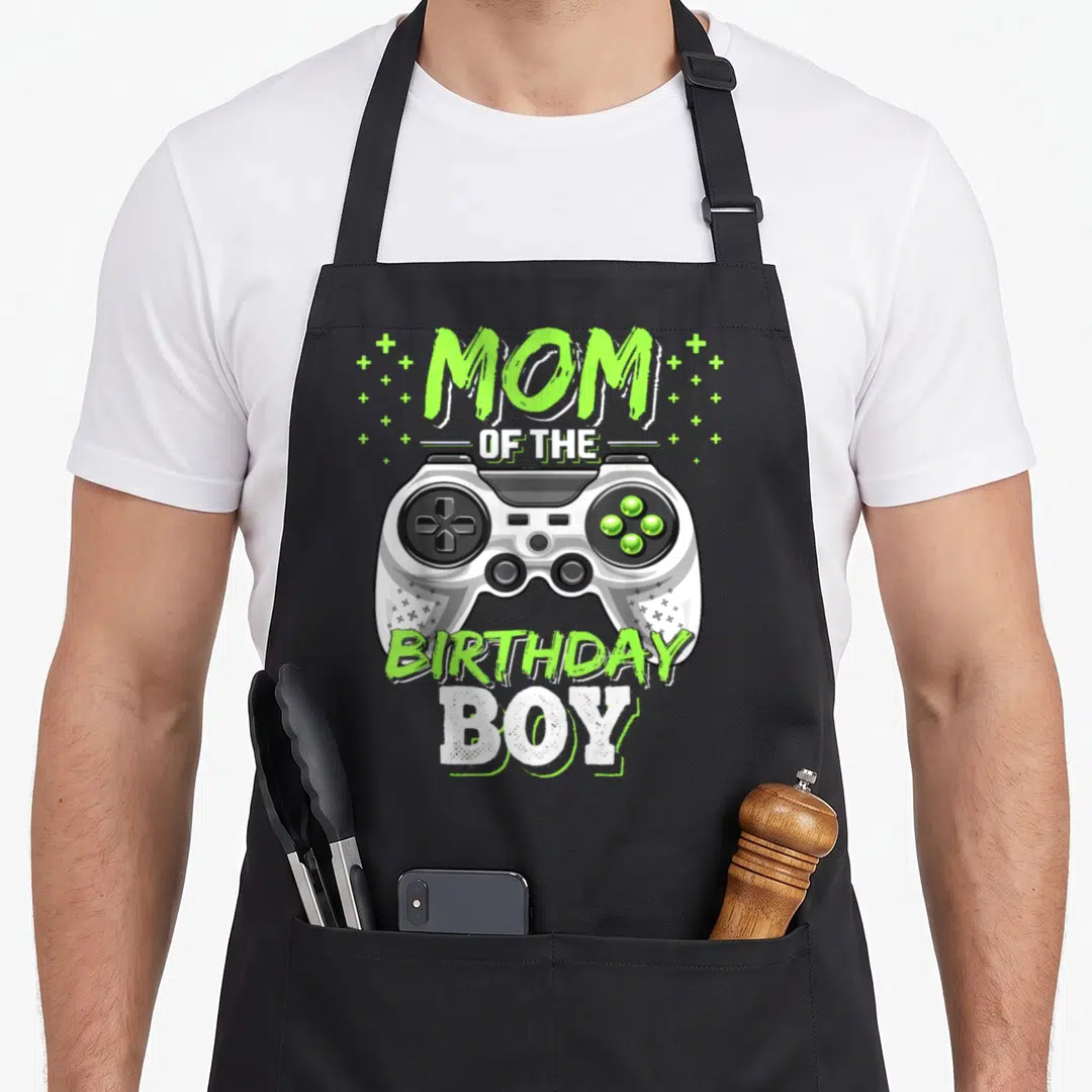 Video birthday apron - MOM OF THE BIRTHDAY BOY text above a white game controller with lime green plus signs — shown on a black cotton canvas apron against a white background