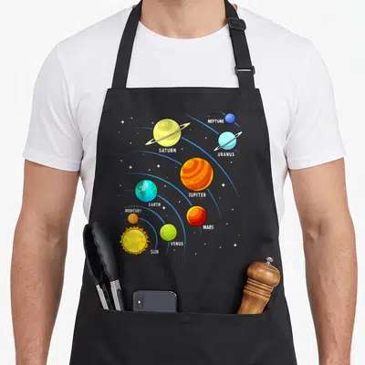 Colorful planet science apron - Cartoon solar system with labeled planets in blue, yellow, orange, red, and green — shown on a black cotton canvas apron against a white background