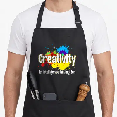 Drawing creative apron - White text Creativity over blue red yellow purple white paint splatters — shown on a black cotton canvas apron against a white background