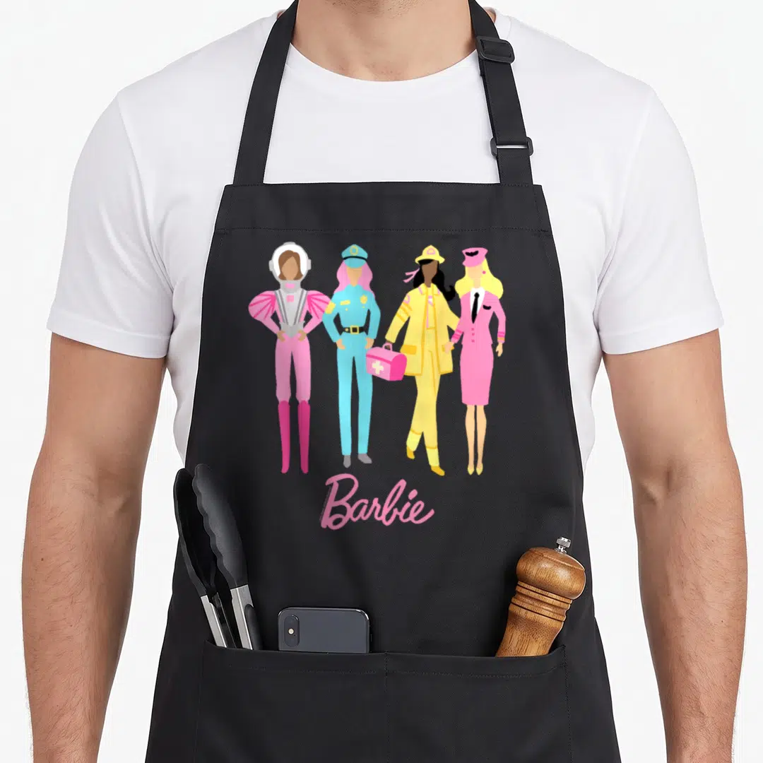 Anniversary 60th apron - Four women in astronaut, police, medical, and pilot outfits in pink blue yellow with Barbie — shown on a black cotton canvas apron against a white background