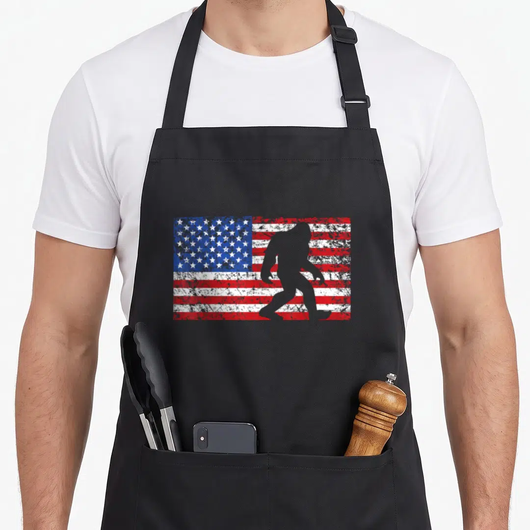 Flag bigfoot 4th of july apron - Grey Bigfoot silhouette walking over a distressed red white and blue American flag — shown on a black cotton canvas apron against a white background