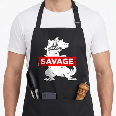 Reptar savage apron - White cartoon monster with red SAVANGE banner on black apron — shown on a black cotton canvas apron against a white background
