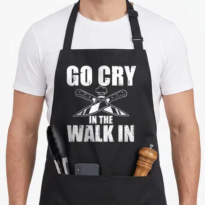 Funny culinary aprons - White distressed text Go Cry In The Walk In above crossed knives and chef hat — shown on a black cotton canvas apron against a white background