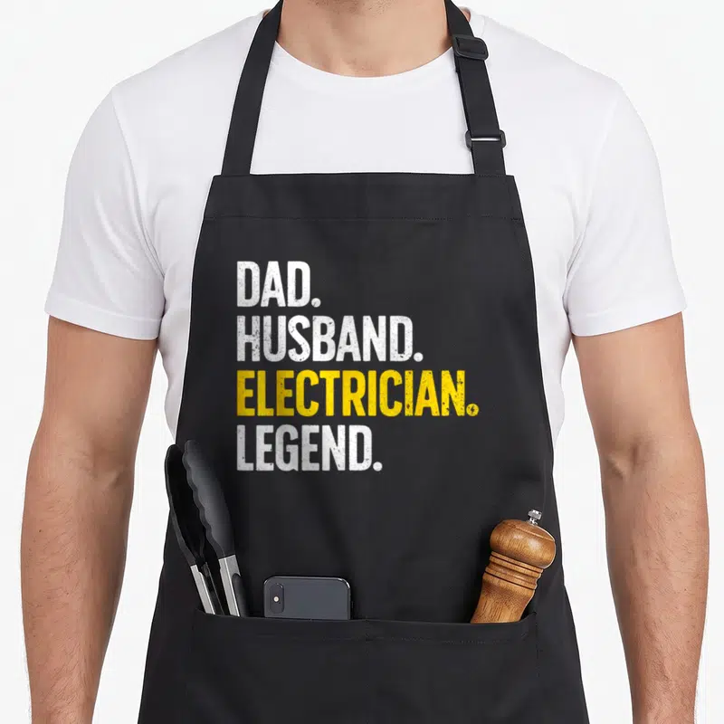 Husband electrician apron - Stacked white and yellow distressed text reading DAD HUSBAND ELECTRICIAN LEGEND on black — shown on a black cotton canvas apron against a white background