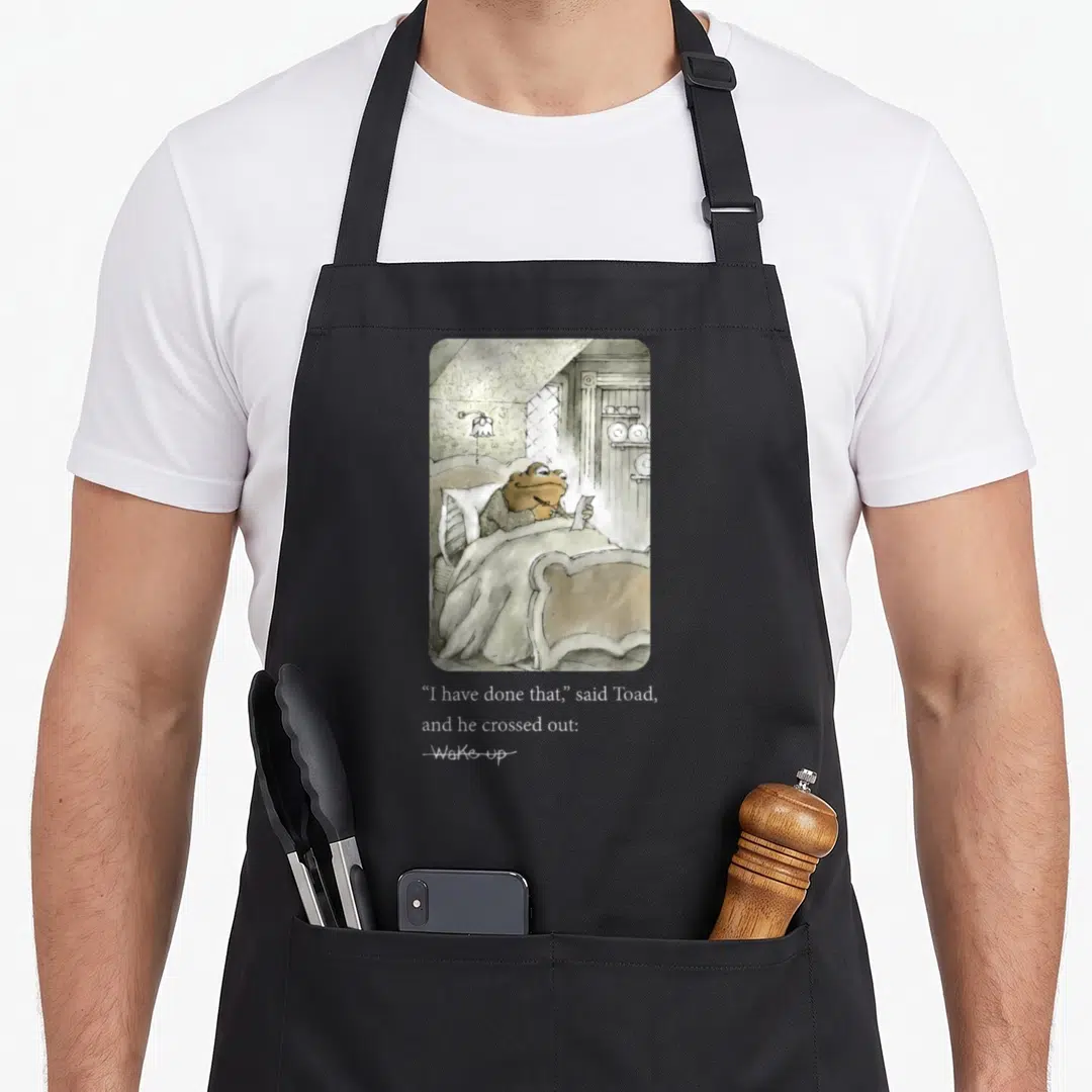 Toad frog apron - Watercolor toad in bed with text I have done that said Toad crossed out Wake up — shown on a black cotton canvas apron against a white background