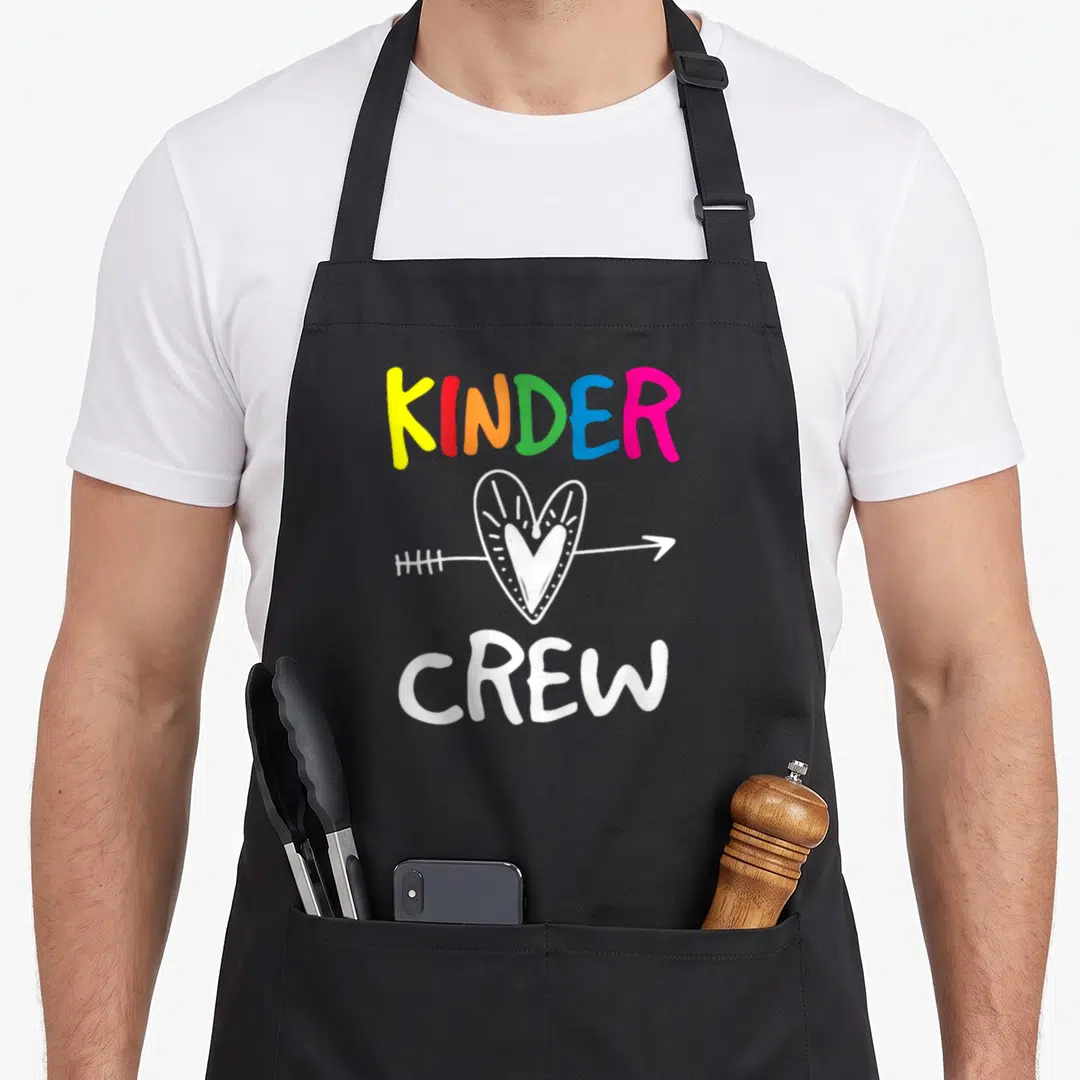 Kinder crew kindergarten teacher apron - Rainbow KINDER CREW text above white heart pierced by arrow — shown on a black cotton canvas apron against a white background