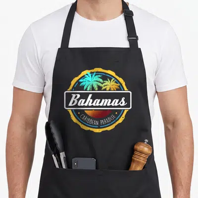 Proud bahamian apron - Circular emblem with two palm trees, sunset, and text Bahamas and CARIBBEAN PARADISE — shown on a black cotton canvas apron against a white background