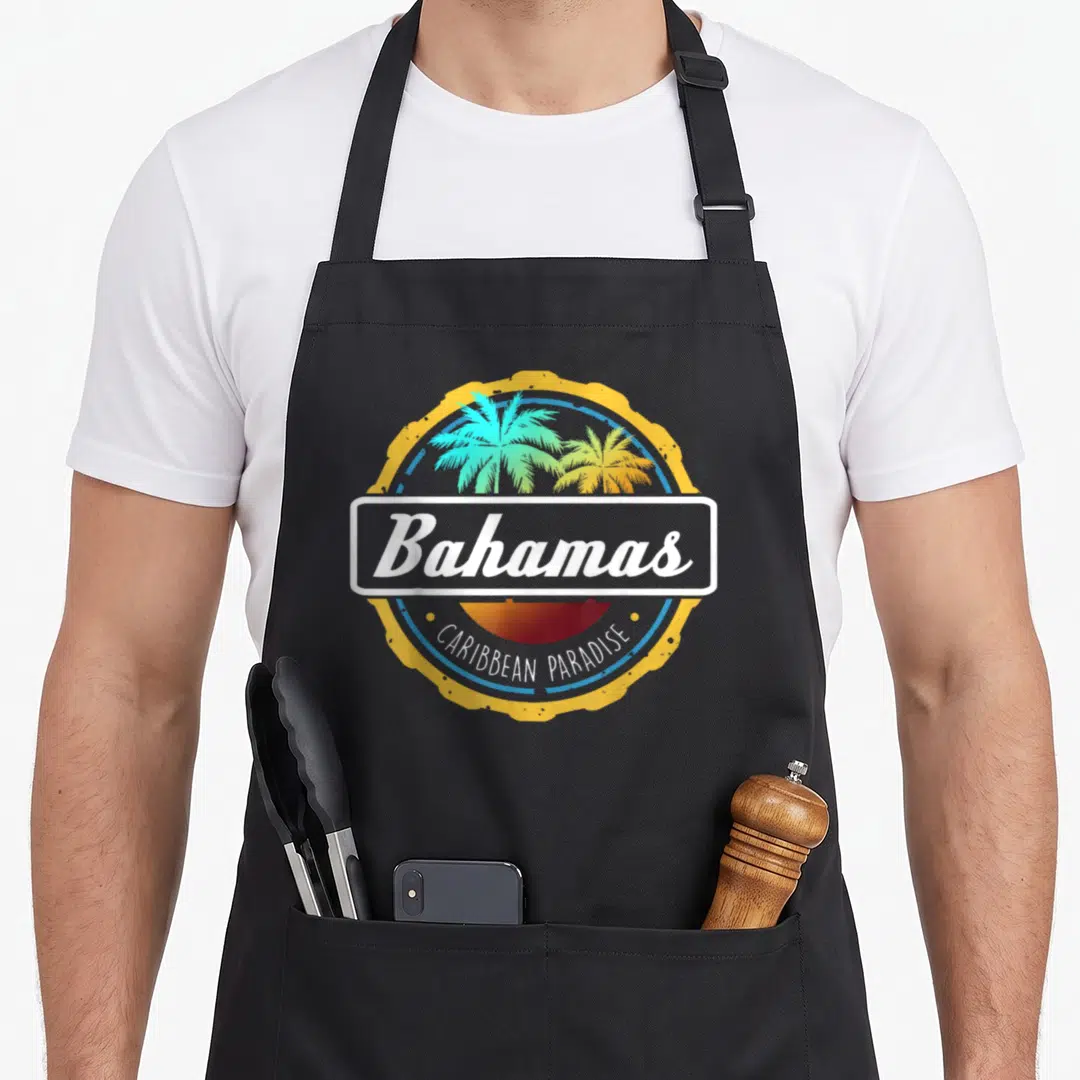 Proud bahamian apron - Circular emblem with two palm trees, sunset, and text Bahamas and CARIBBEAN PARADISE — shown on a black cotton canvas apron against a white background